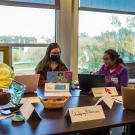 Gathered around a table with windows behind them looking out on trees and a grassy field, Edu Nguema, Rudo Chasi, a female student in a mask, Sushila Thing and a male student in a mask each have a laptop in front of them. Placards on the table identify this group as \"Developing B Nations\" and some of the countries they represent as Fiji and Nepal.